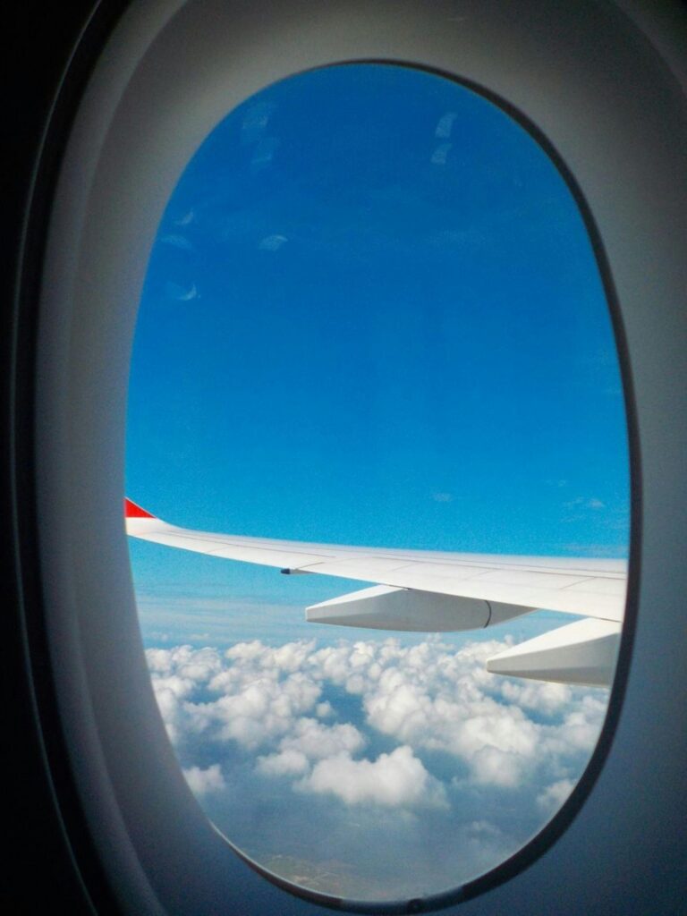 View of an airplane wing and fluffy clouds through a plane window, with a bright blue sky.
